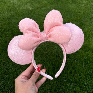 May include: A pink sequined headband with large mouse ears and a bow. The ears and bow are made of a soft, textured material. The headband is held by a hand with red nail polish against a green background.
