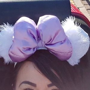 May include: A lavender satin bow headband with white, fluffy ear accents. The bow is large and centered, with a sparkly band. The headband is a fun accessory, perfect for theme parks or special events.