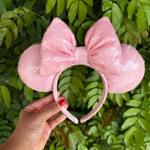 May include: Pink sequined Minnie Mouse ears headband with a large bow. The headband is made of a soft, flexible material and is perfect for any Disney fan.