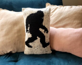 May include: Three decorative pillows on a blue couch. The center pillow is white with a black silhouette of a Bigfoot. The pillow on the left is beige with a brown grid pattern. The pillow on the right is pink.
