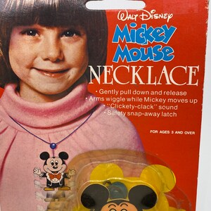 May include: A red and white cardboard package featuring a girl wearing a necklace with a Mickey Mouse pendant. The package reads "Walt Disney Mickey Mouse Necklace" and includes instructions for use. The instructions state "Gently pull down and release Arms wiggle while Mickey moves up "Clickety-clack" sound Safety snap-away latch" and "For ages 3 and over".