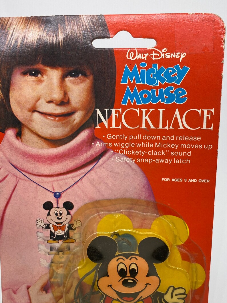 May include: A red and white cardboard package featuring a girl wearing a necklace with a Mickey Mouse pendant. The package reads "Walt Disney Mickey Mouse Necklace" and includes instructions for use. The instructions state "Gently pull down and release Arms wiggle while Mickey moves up "Clickety-clack" sound Safety snap-away latch" and "For ages 3 and over".