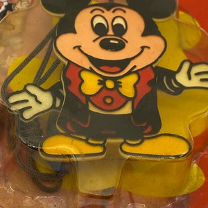 May include: A plastic Mickey Mouse character wearing a tuxedo with a yellow bow tie. The character is attached to a black string and is packaged in a clear plastic bag.