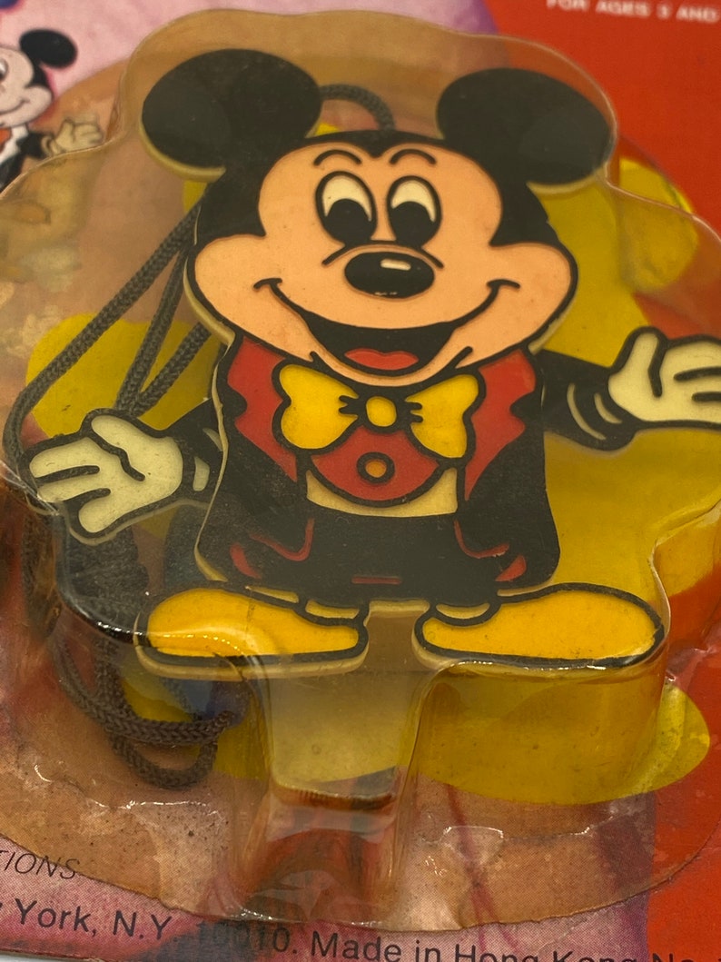 May include: A plastic Mickey Mouse character wearing a tuxedo with a yellow bow tie. The character is attached to a black string and is packaged in a clear plastic bag.
