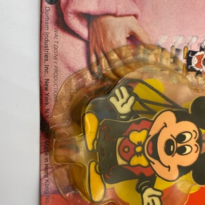 May include: A plastic Mickey Mouse character keychain with a yellow background. The keychain is packaged in a clear plastic blister pack with a red, white and yellow cardboard backing. The cardboard backing has the text "WALT DISNEY PRODUCTIONS" and "Durham Industries, Inc., New York, N.Y. 10010. Made in Hong Kong No. 1562."