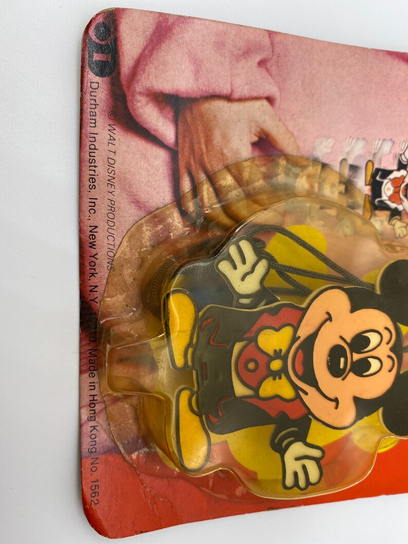 May include: A plastic Mickey Mouse character keychain with a yellow background. The keychain is packaged in a clear plastic blister pack with a red, white and yellow cardboard backing. The cardboard backing has the text "WALT DISNEY PRODUCTIONS" and "Durham Industries, Inc., New York, N.Y. 10010. Made in Hong Kong No. 1562."