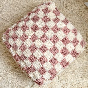 Puede incluir: Un cojín cuadrado, afelpado, con un diseño de tablero de ajedrez en rosa empolvado y crema. El cojín tiene un pelo rizado texturizado y un borde con flecos. El cojín está colocado sobre una alfombra crema texturizada. Mide unos 30 cm x 30 cm.