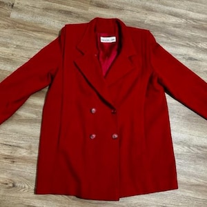 May include: A double-breasted, red blazer with two rows of buttons. The blazer has a classic lapel design and long sleeves. The garment is made of a smooth fabric and is a solid colour. The blazer is a piece of outerwear.