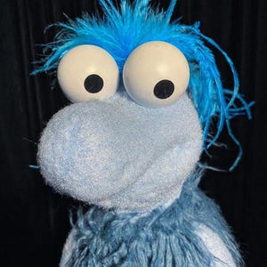 May include: A blue puppet featuring a fuzzy, light blue head and a textured, darker blue body. The puppet has two large, round white eyes with black pupils and a shock of bright blue hair on top. The material appears soft and plush throughout.