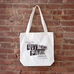 May include: White tote bag with black text that reads "Greetings From... Ditmas Park Brooklyn The Pride of Flatbush." The bag has two white handles and is hanging against a red brick wall.