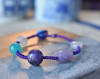 May include: A handmade bracelet featuring a variety of gemstone beads in shades of purple, blue, and clear. The beads are strung on a purple cord. The bracelet is displayed on a gray surface.