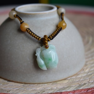 May include: A jade pendant necklace with a light green and white carved floral design. The necklace has a black and gold braided cord with yellow and white beads. The pendant is displayed on a small, light gray ceramic bowl.