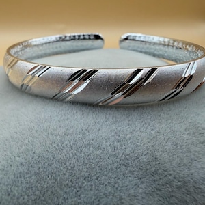 Pode incluir: Uma pulseira de bracelete prateada com um acabamento fosco texturizado. A pulseira apresenta listras diagonais polidas em sua superfície. O design aberto facilita o uso. Esta joia é adequada para o uso diário.