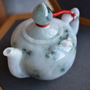 Könnte beinhalten: Kleine Teekanne aus Jade, weiß mit grünen Adern, mit Deckel und Ausguss. Roter Kordeldetail am Deckel. Auf dunkler Oberfläche platziert.