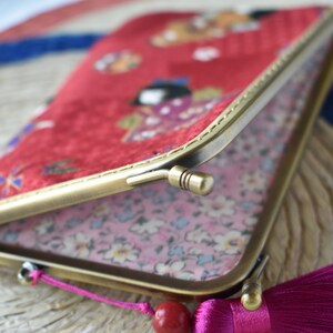 May include: A red clutch with a gold-colored frame and a pink tassel. The clutch features a Japanese-style pattern with figures and floral designs. The interior lining is a light pink floral print.