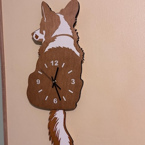 Può includere: Un orologio da parete a forma di cane seduto con motivi di pelo marrone e bianco. Il quadrante è integrato nel corpo del cane, con numeri bianchi nelle posizioni 12, 3, 6 e 9, insieme a lancette nere per ore, minuti e secondi.