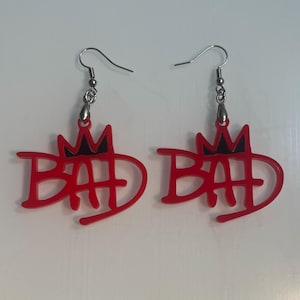 May include: A pair of red acrylic earrings with the word "BAD" in a stylized font, topped with a black crown design. The earrings have silver-colored fish hook ear wires.