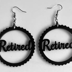 May include: A pair of black earrings with a circular design. The earrings feature a gear-like outer ring and the word "Retired" in a cursive font within the circle. The earrings hang from silver-colored fish hook ear wires.