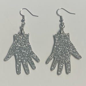 May include: A pair of silver glitter hand-shaped earrings. Each earring features a hand silhouette with outstretched fingers, suspended from a silver fish hook ear wire. The earrings are designed to catch the light and sparkle.