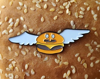 Könnte beinhalten: Ein burgerförmiger Emaille-Pin mit einem Cartoon-Gesicht, Schnurrbart und weißen Flügeln, auf einem Sesambrötchen. Der Pin ist orange, gelb und schwarz. Das Brötchen ist goldbraun.