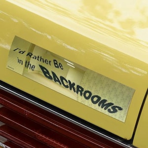 May include: A yellow car bumper with a sticker that reads "I'd Rather Be in the BACKROOMS" in black, pixelated font. The sticker is rectangular and has a blurred background.