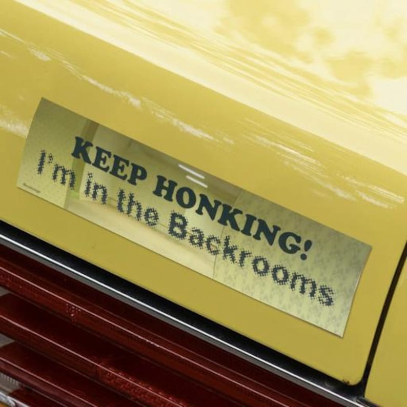 May include: A rectangular bumper sticker with the text "KEEP HONKING! I'm in the Backrooms" in dark green font. The sticker is affixed to a yellow car, with a red and chrome trim visible below.