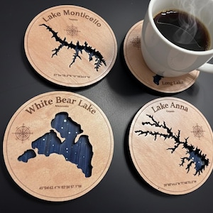 May include: Wooden coasters with lake outlines, including Lake Monticello, White Bear Lake, and Lake Anna. Each coaster features a compass rose design and lake name. A white mug with coffee sits atop a coaster. The coasters are made of light-colored wood.
