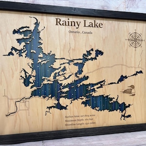 May include: Framed wooden map of Rainy Lake, Ontario, Canada. The laser-cut map features a blue and green lake design, a compass rose, and a loon illustration. The text includes the lake's surface area, maximum depth of 109.73 metres, and shoreline length.