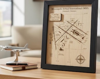 May include: A framed wooden map of the Minneapolis-St Paul International Airport (MSP) in Minnesota. The light wood surface features a dark engraved runway layout, a compass rose, and a small rectangular plaque with text including the date.