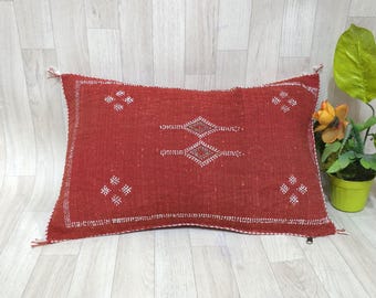 May include: A rectangular, red pillow with white geometric patterns. The pillow has a textured appearance and is adorned with tassels on the corners. A small green potted plant with yellow flowers is next to the pillow.