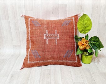 May include: A square, rust-colored throw pillow featuring a white embroidered border and a central white geometric motif. The pillow is accented with small, light blue embroidered leaf-like patterns in each corner, showcasing a textured fabric finish.