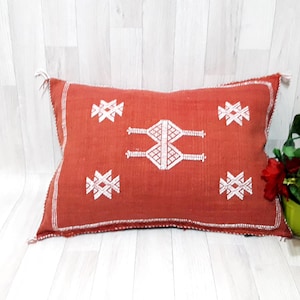 May include: A red rectangular pillow with white embroidered geometric patterns. The pillow is on a white wood floor.