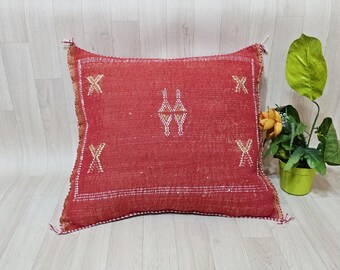 May include: A square, red decorative pillow with white embroidered geometric patterns. The pillow has a textured appearance and is adorned with tassels on the corners. A small plant in a green pot is to the right of the pillow.