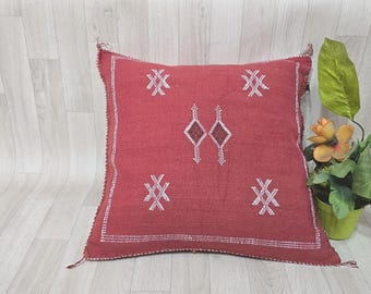 May include: A red square throw pillow with white embroidered geometric patterns. The pillow is on a light wood floor.