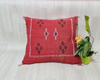Könnte beinhalten: Ein quadratisches rotes Zierkissen mit weißem Zierstichrand und schwarzen rautenförmigen geometrischen Mustern. Das Kissen hat eine strukturierte Stoffoberfläche mit sichtbaren Nähten und steht neben einer kleinen grünen Topfpflanze.