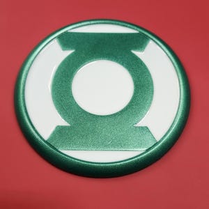 May include: Green Lantern logo emblem. The emblem features a green and white design with a circular shape. The outer ring and the central symbol are green, while the background is white. The emblem is set against a red background.