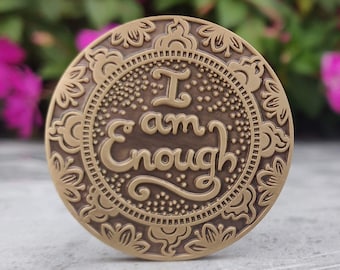 May include: A round, gold-coloured metal token with an ornate design and the words "I am Enough" inscribed in a decorative font.