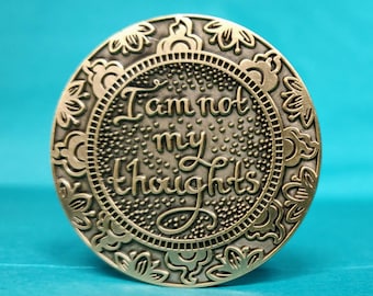 May include: A circular, antique brass-toned coin with the words "I am not my thoughts" inscribed in a decorative font. The coin has a floral border and a dotted pattern around the text.