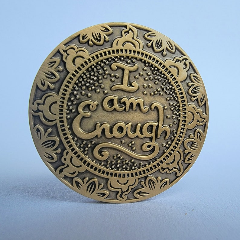 Pu&ograve; includere: Una moneta di metallo dorato con un design ornamentale attorno al bordo. Il centro della moneta presenta le parole "I am Enough" in una scrittura decorativa.