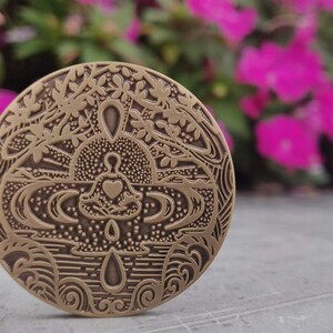 Pu&ograve; includere: Un medaglione rotondo in metallo color ottone con un design intricato. Il design presenta una persona seduta in posizione di loto con un cuore sul petto, circondata da alberi stilizzati, acqua e onde.