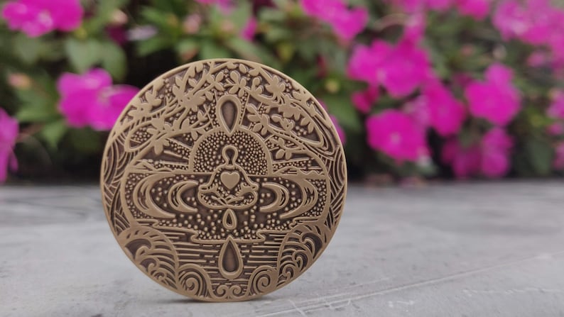 Pu&ograve; includere: Un medaglione rotondo in metallo color ottone con un design intricato. Il design presenta una persona seduta in posizione di loto con un cuore sul petto, circondata da alberi stilizzati, acqua e onde.