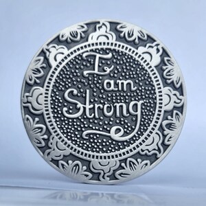 May include: A round silver-toned metal coaster with an ornate floral design around the edge. The centre features a black background with the words "I am Strong" in a stylised font.