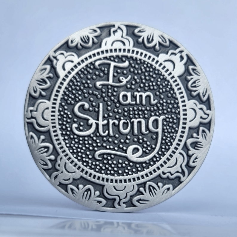 May include: A round silver-toned metal coaster with an ornate floral design around the edge. The centre features a black background with the words "I am Strong" in a stylised font.