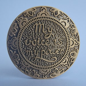 Pu&ograve; includere: Un medaglione rotondo in metallo dorato con un design ornamentale e le parole "My voice is my power" in un carattere decorativo.