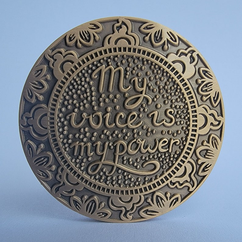 Pu&ograve; includere: Un medaglione rotondo in metallo dorato con un design ornamentale e le parole "My voice is my power" in un carattere decorativo.