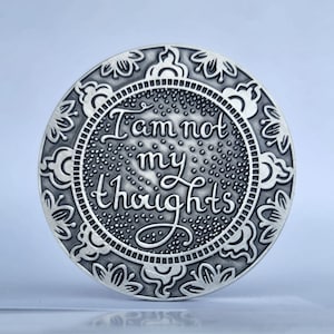 May include: A silver round coin with an ornate border and the words "I am not my thoughts" inscribed in the centre.