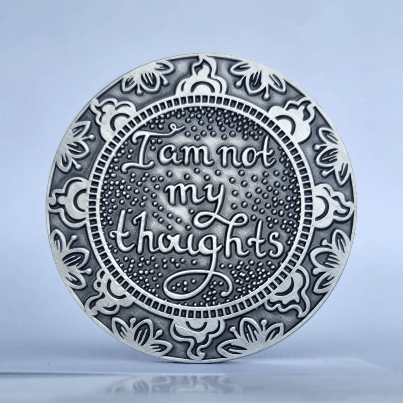 May include: A silver round coin with an ornate border and the words "I am not my thoughts" inscribed in the centre.