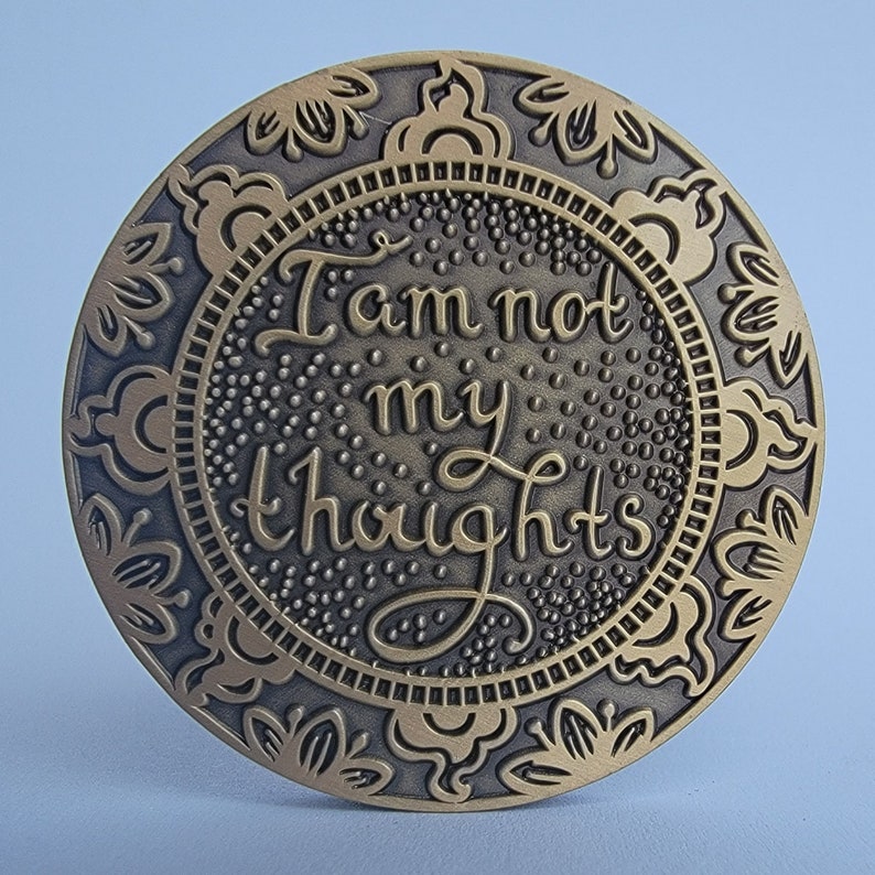 Pu&ograve; includere: Un medaglione rotondo in metallo color ottone con un design ornamentale. Il centro del medaglione presenta il testo "Io non sono i miei pensieri" in un carattere decorativo.