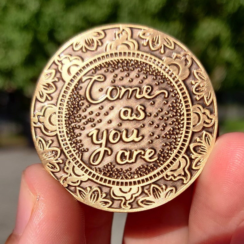 Pu&ograve; includere: Una moneta di metallo color oro con un designo ornato e le parole "Come as you are" incise su di essa.