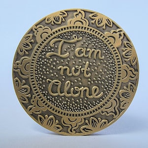 Pu&ograve; includere: Un gettone rotondo in metallo dorato con un design ornamentale. Il centro del gettone presenta le parole "Non sono solo" in un font stilizzato.